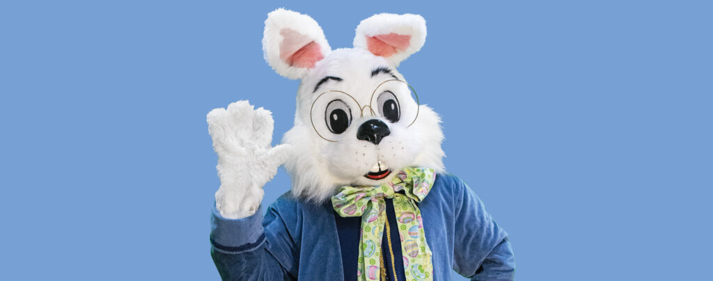 White bunny costume with glasses and a colorful bow tie waving for photos with Easter Bunny, set against a solid blue background